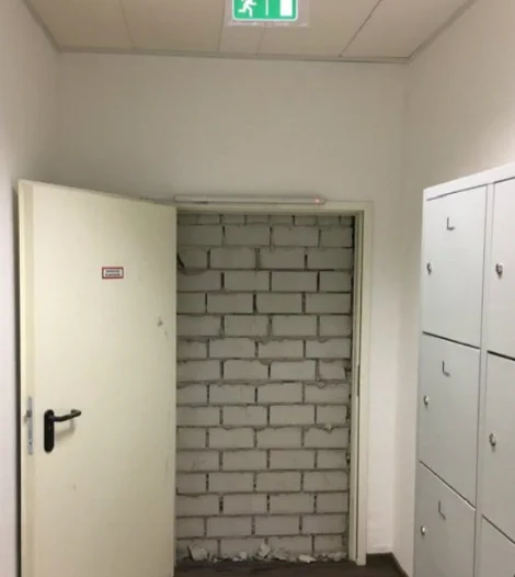Ein Flur mit einer weißen Feuerschutztür, darüber ein grünes Schild mit Notausgang. Leider kann man nicht durch weil alles zugemauert ist. 