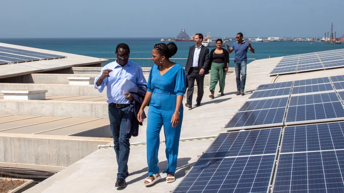 Five people walking on a roof top next to solar panels, ocean in the background