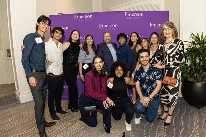 A group of people smile and pose in front of a purple background with the words "Emerson College."