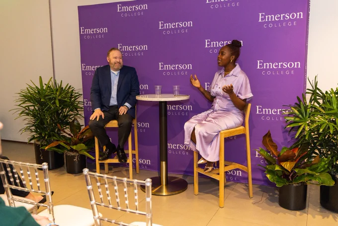 Two people sit at a table top and engage in conversation in front of a purple background with the words "Emerson College."