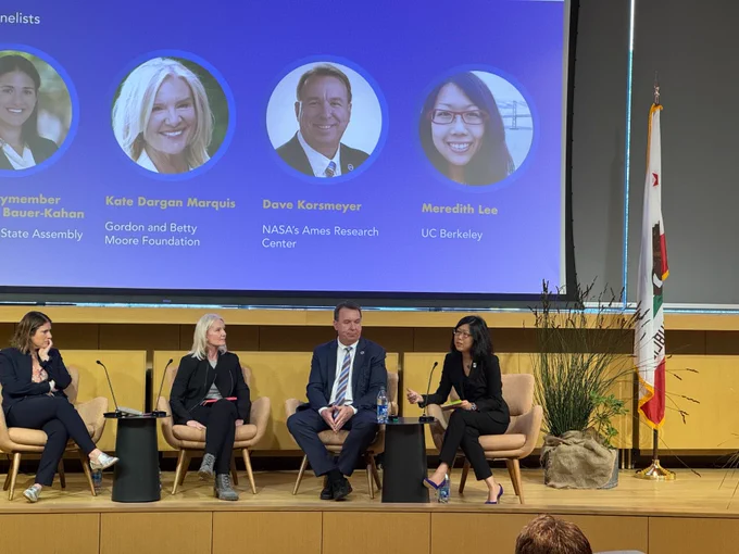 A photo of the panel on a stage with a California flag and the projector screen behind them showing the panelists photos and names.