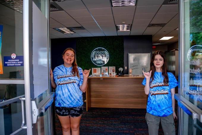 Two students stand at the entrance of their storefront. The organization is Roadrunner Productions, and they are wearing Tie Dye shirts read the organization name.