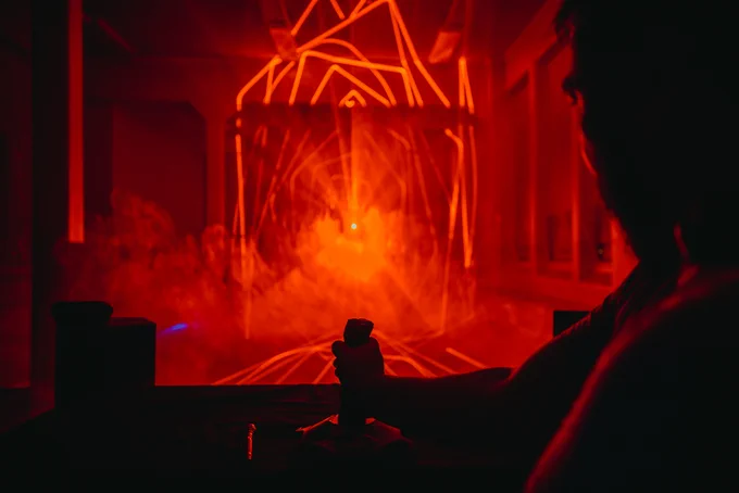 A person stands to the right of the frame, manoeuvring a joystick. The joystick controls the laser that fills most of the screen, currently coloured a vivid orange-red and casting geometric patterns out across the room.