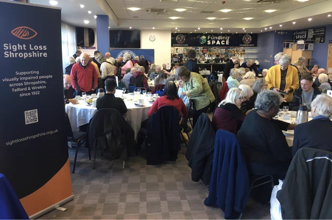 An image of the busy event. Lots of people sat and stood around tables with the Sight Loss Shropshire sign at the forefront.