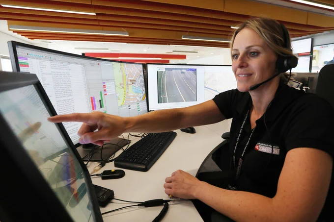 A female fire control operator, with 3 computer screens behind her is pointing towards another screen.