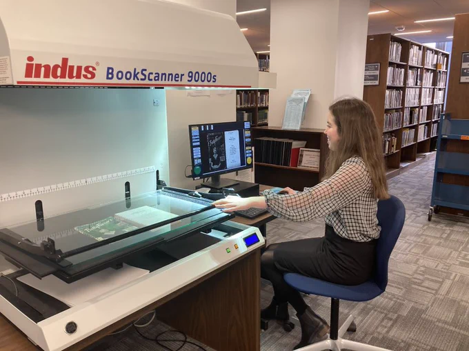 A woman sitting on an task chair at a large scale book scanner. She is operating the scanner at a desktop computer at the State Library of North Carolina. 