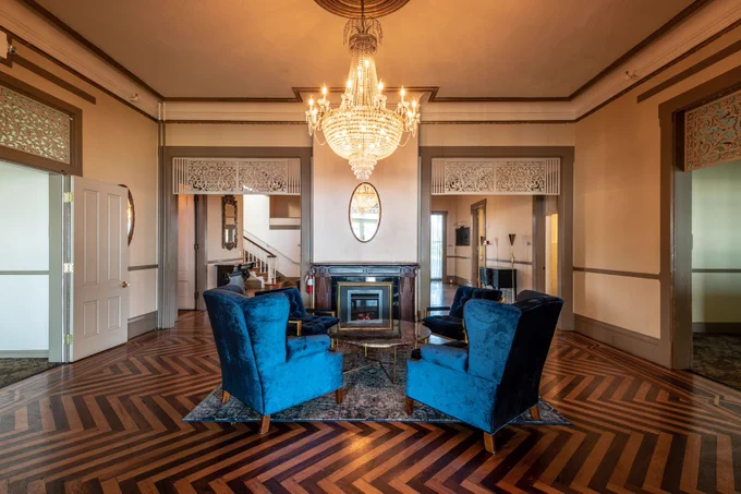 An image of a foyer in a historic mansion with lounge sofa chairs, a fireplace, and a chandelier. 