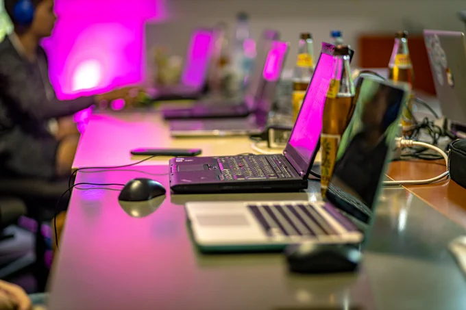 Eine lange Tischreihe mit Laptops, von der Seite gesehen. Auf den Tischen sieht man Computermäuse und Flaschen mit Mate-Tee. Im Hintergrund arbeitet ein Mensch mit Kopfhörern an einem Laptop. Die Szene ist pink beleuchtet.
