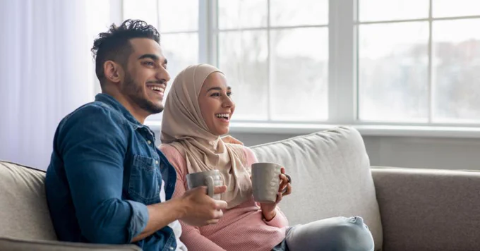 A couple sits on a couch in front of a brightly lit window with mugs. The woman wears a hijab.