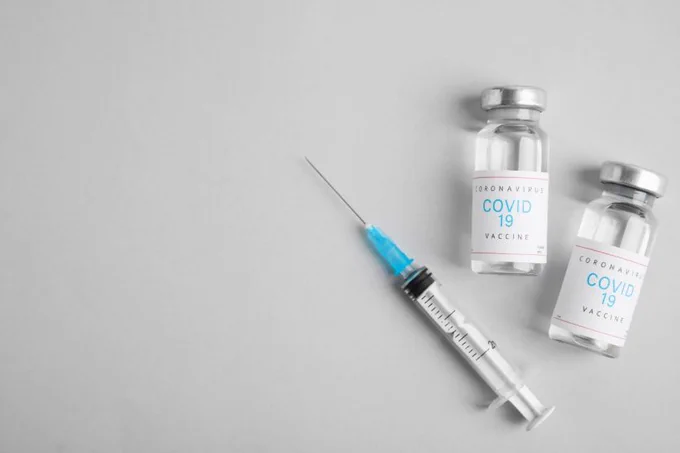 L'image est une photographie avec un fond gris et sur la droite de celle-ci se trouve le nécessaire pour réaliser un vaccin, à savoir une seringue et deux flacons de vaccins contre le Covid-19.