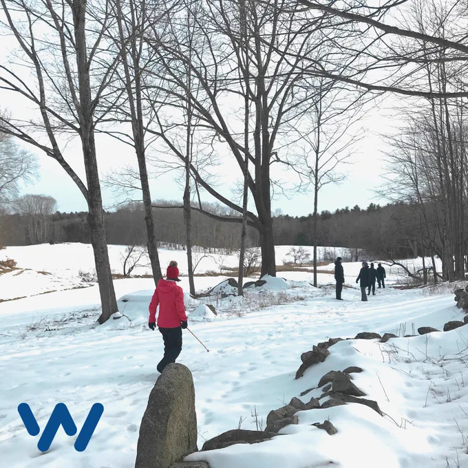 A young woman wearing snow gear walking along a snowy path using a sighted cane. Others are ahead of her and a field of snow with bare winter trees are in the background.
