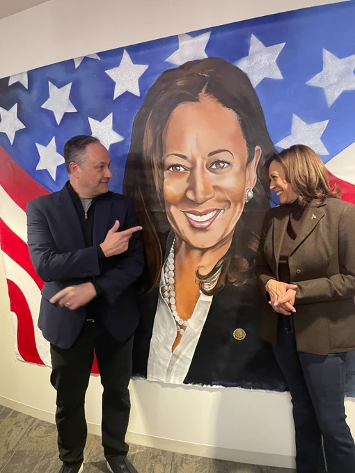 Vice President Kamala Harris and Second Gentleman Douglas Emhoff posing for a picture in front of wall art of Vice President Kamala Harris at Biden-Harris campaign headquarters