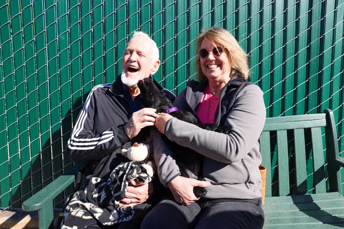 In this photo, a man and woman sit on a bench and hold their black Lab puppy.