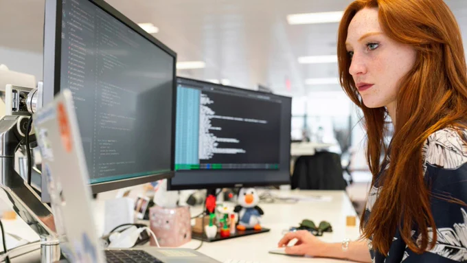 Une femme dans son bureau face à deux écrans d'ordinateur.