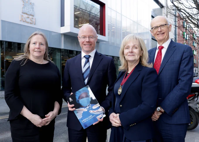 
A group of four people stand in front of an RCSI building. All are smiling. The man in the middle, Ireland’s Minister for Health Stephen Donnelly holds a document in his hands with the words ‘Surgical Hubs: Clinical Guidance’ on it. 
