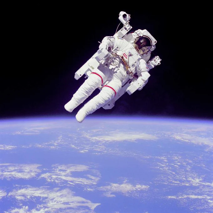 An astronaut floats in space while attached to a chair-like device. The Earth is visible in the background.