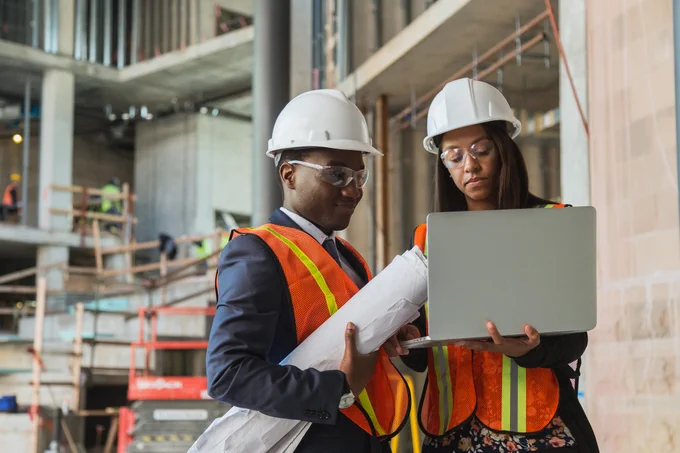 A male and female engineer wearing hard hats are looking at a computer in a construction site.