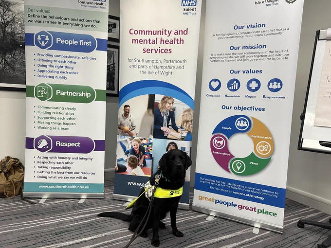Guide dog Loney  sat in front of three banners showcasing. The three organisations involved in the conference. She’s wearing her harness.