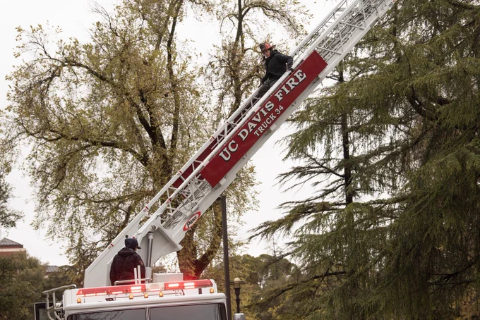One UCDFD Firefighter coming down the latter of a rig that says ‘UC Davis Fire Truck 34’, with gloomy sky and trees behind him. Another UCDFD Firefighter at the start of the latter is shown providing assistance.