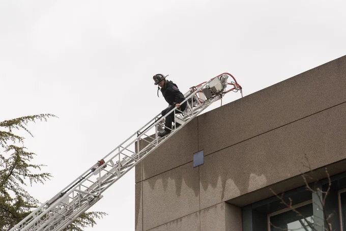A UCDFD Firefighter coming down from the top of latter of Truck 34 over a wet building and a cloudy sky in the background.