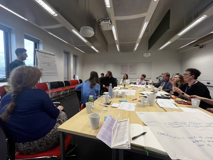 Around 12 people sitting at a table with lots of post it notes and flipchart paper watching Callum present