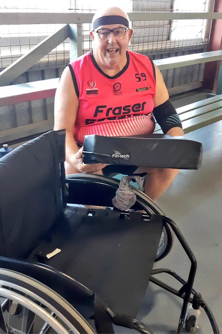 This is me, a smiling older male wearing a pink singlet and black headband seated on a wooden bench holding my Invacare cushion. My basketball wheelchair is in the foreground 