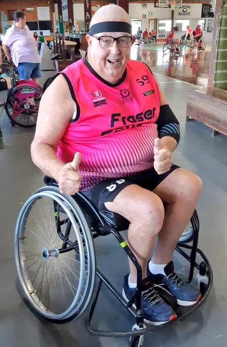 This is me, a smiling older male wearing a pink singlet and black headband seated in my basketball wheelchair with both thumbs up 