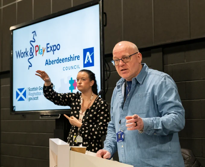 Photograph courtesy of Aberdeenshire Council. Ewen Allardyce addresses the audience from behind a lecturn at the Employment Support Team's Work & Play Expo Event.  The lady beside Ewen is using sign language and the display screen shows the Work & Play Expo branding and three logos.