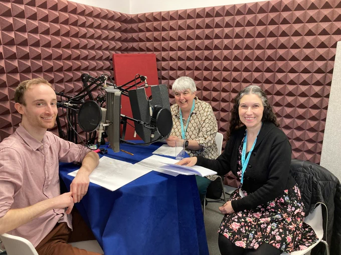 Photo of three people sitting at a circular table with microphones in a recording studio.