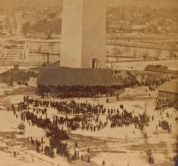 Historic black and white photo of hundreds of people walking around the paths and lawns around the base of the white stone tower of the Washington Monument on a snowy day.