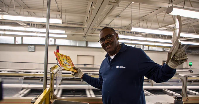 Man wearing a long-sleeved blue Toronto Public Library shit. He has his arms outstretched. In each hand, he holds a book. He's standing in front of the library's book sorter. 