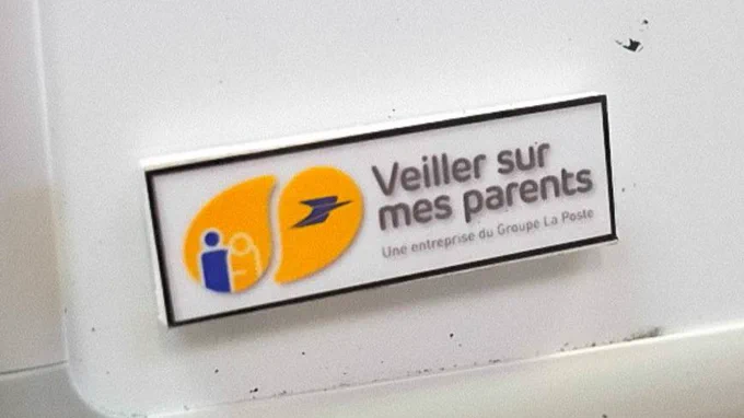 Sur un mur blanc est accroché un logo sur lequel est écrit « Veiller sur les parents » et en plus petit « Une entreprise du Groupe La Poste ».