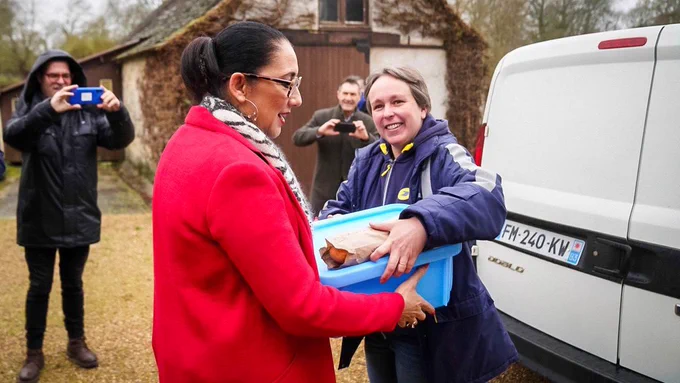 Nadège, factrice participant aux « services seniors » de La Poste tend un panier repas à la ministre. À droite de l’image se trouve la camionnette de La Poste. 

En arrière-plan, on distingue une maison et deux personnes prenant des photos.