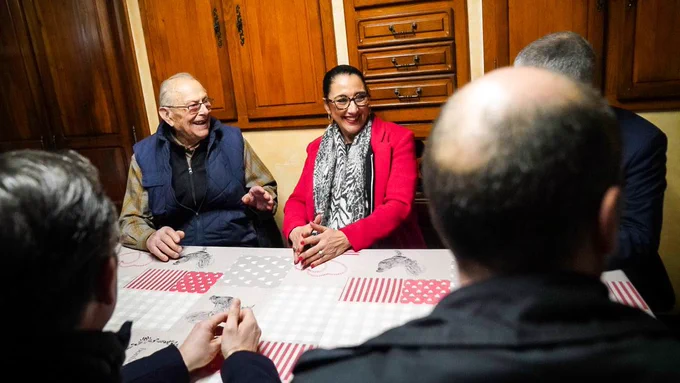 Fadila Khattabi est attablée aux côtés de René, bénéficiaire des « services seniors » de La Poste. Face à eux se trouvent deux personnes de dos, également attablées.

En arrière-plan, on distingue des meubles en bois.