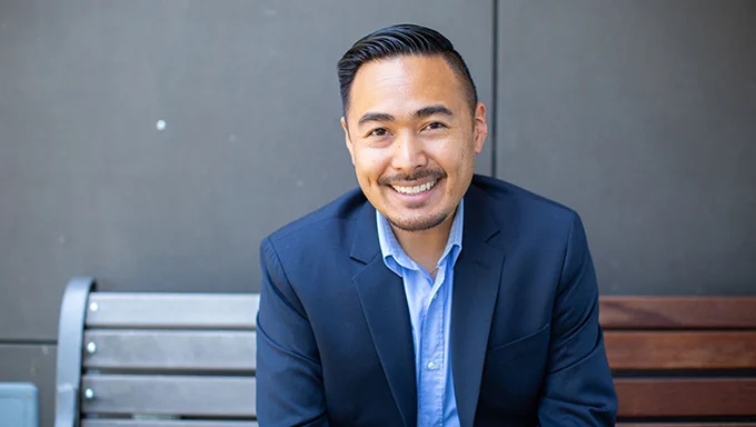 Lecturer Justin Virrey sits on a wooden bench outside and smiles, wearing a navy blazer and light blue button down shirt.