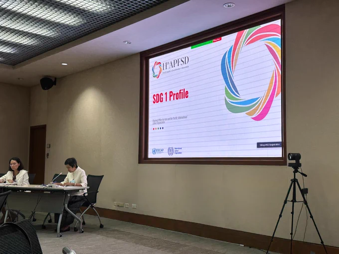 Two women sit in the far left corner of a large room, in front of a projector that reads 'SDG 1 Profile, 11th APFSD'. There is a camera filming the room.