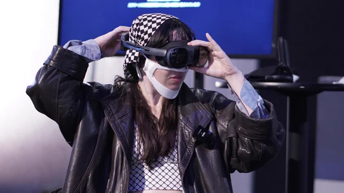 Nat, a white non-binary person with dark brown long hair and step bangs. They are wearing a leather jacket with black and white checkered shirt under and a matching checkered head band. They are adjusting the VR headset they are wearing.
