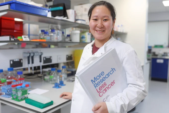 A woman in a laboratory wearing a white coat and holding a folder with the words "More Research, Less Cancer" on it.