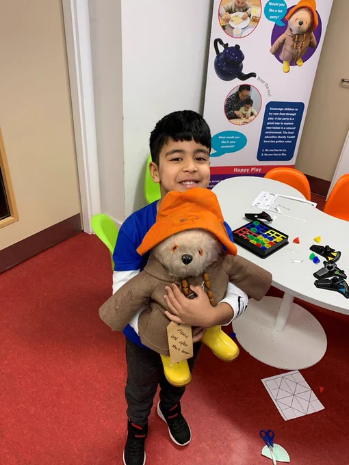 Young boy smiling and holding a Paddington Bear teddy. He is stood in front of a table with a game spread across it. At the back is a banner that features Paddington Bear.