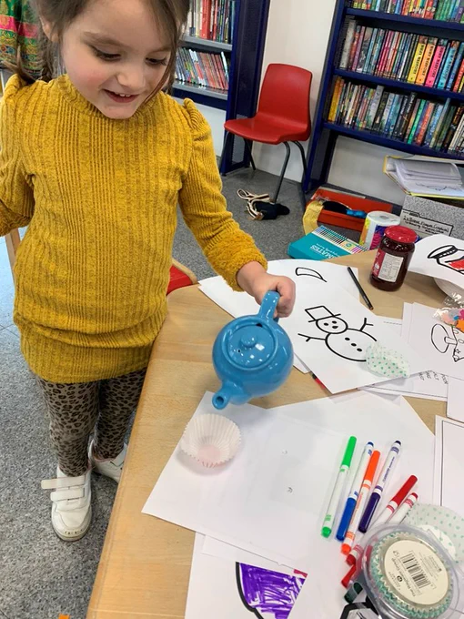 A young girl is pouring imaginary tea from a teapot into a cupcake case. She is stood at a table covered in colouring activities and pens. 