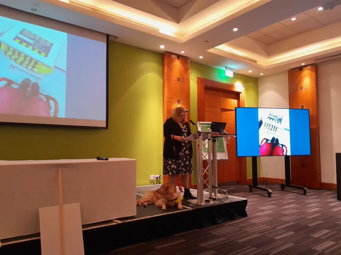 Photo of Amy, 5 foot 2 female with blonde hair wearing a bright flowery dress and black cardigan and black glasses. Next to her guide dog Ava, yellow lab. On the lecturn with a PowerPoint presenting showing a picture of the lower half of a person wearing pink trousers and shoes in a wheelchair trying to access a pedal only accessed bin with a sign on top saying offensive waste. 