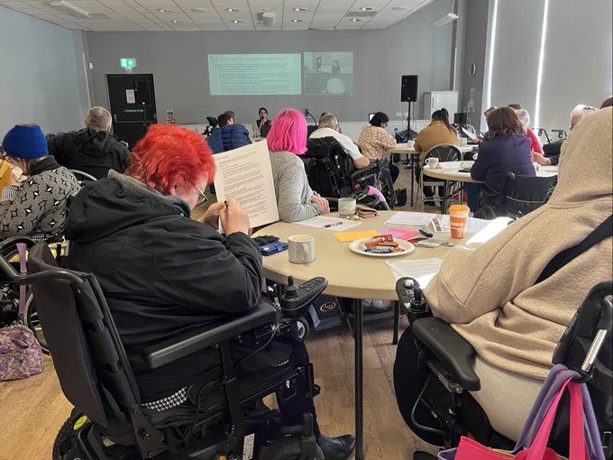 GDA members are gathered around 3 round tables while listening and looking towards a presentation projected at the far end of the room. There are 3 members at the table at the foreground of the image, one with pink hair and one with red hair. The member with red hair is taking notes on their briefing pack.