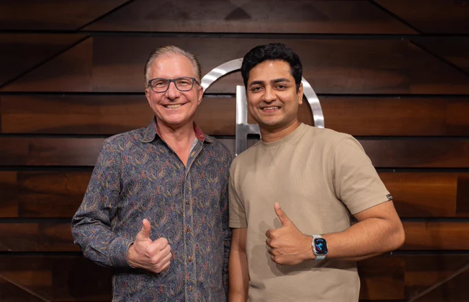 Joz and Kenny Sebastian standing next to each other on stage, against a dark brown wood wall. Kenny is wearing an Apple Watch. 