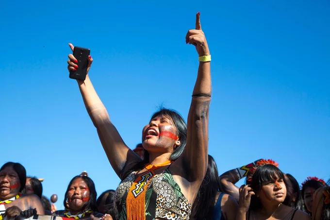 Indigenous woman grips a cell phone and raises her hands to the sky, surrounded by other woman.