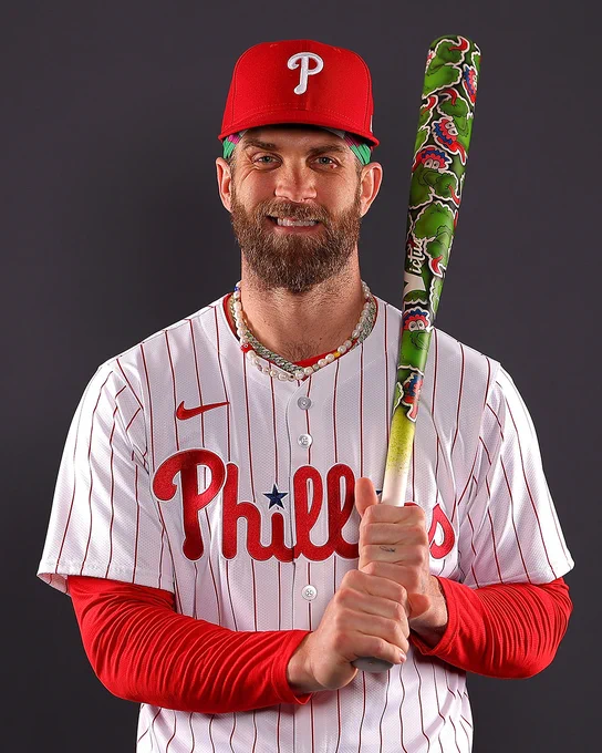 Bryce Harper posing for a portrait in his Phillies uniform during photo day. He's holding a Phanatic-themed baseball bat.