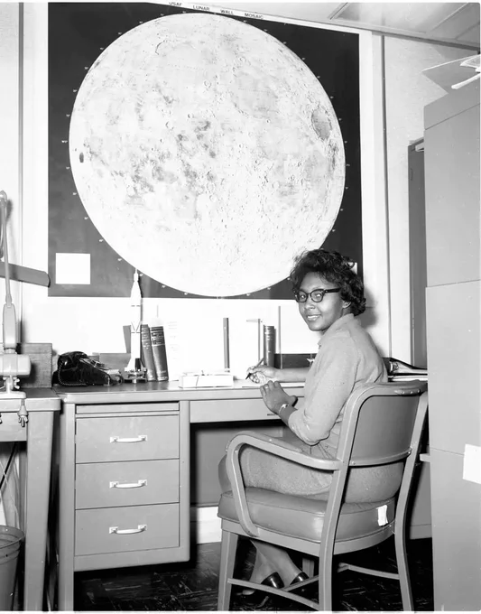 In this black and white image, Jeanette A. Scissum sits at a desk writing on a piece of paper. A photograph of the Moon hangs on the wall behind her. 