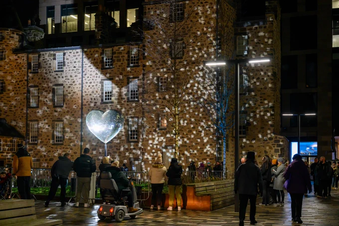 A group of people outdoors looks at an illuminated giant disco ball in a shape of a heart.