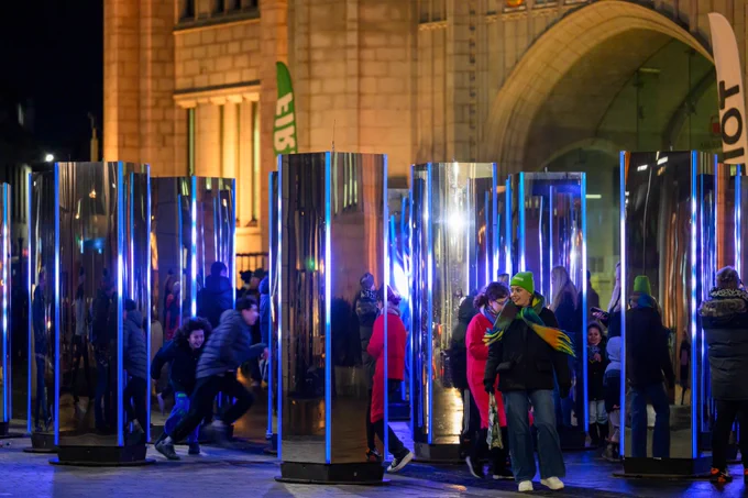 People walking around and looking at an outdoors arts installation featuring tall four-sided mirror panels.
