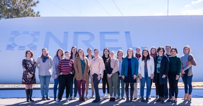 A group of women stand in front of an NREL branded wind turbine.