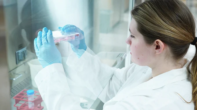 Marie Johns, wearing her lab coat and gloves, looks at a samples she is holding inside a biosafety cabinet.